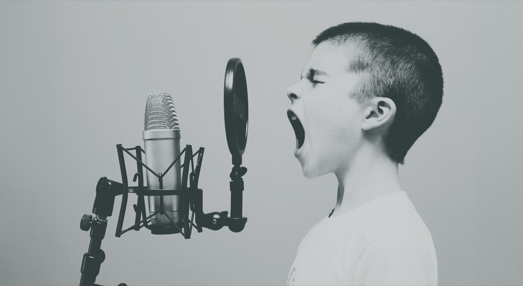 Et svart-hvitt bilde av en ung gutt i en hvit t-skjorte, profilvisning, synger eller roper lidenskapelig inn i en profesjonell studiomikrofon. Intensiteten i uttrykket hans formidler sterke følelser eller en kraftig vokalytelse.